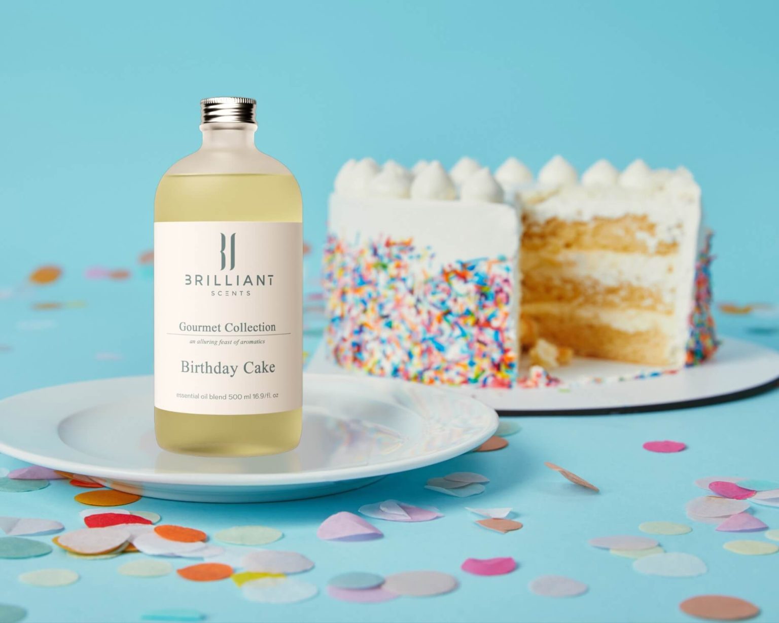 Bottle of Birthday Cake fragrance oil beside a slice of vanilla cake with sprinkles and buttercream frosting, on a pale blue background with confetti.